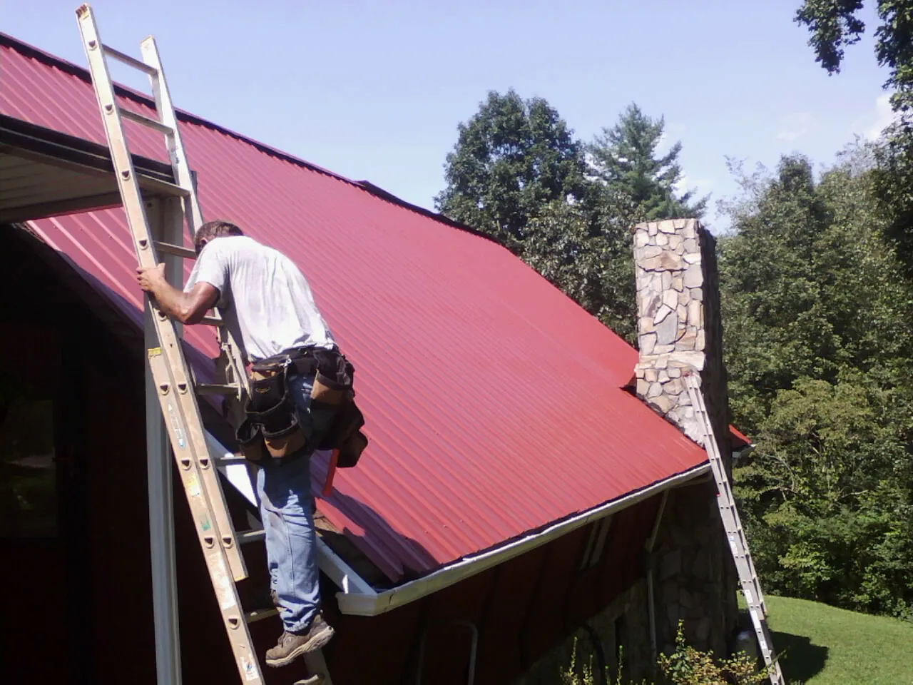 Roofer on a ladder beside a red metal roof and stone chimney — Soffit & Fascia Repair by Roofing Brothers in Manzanita